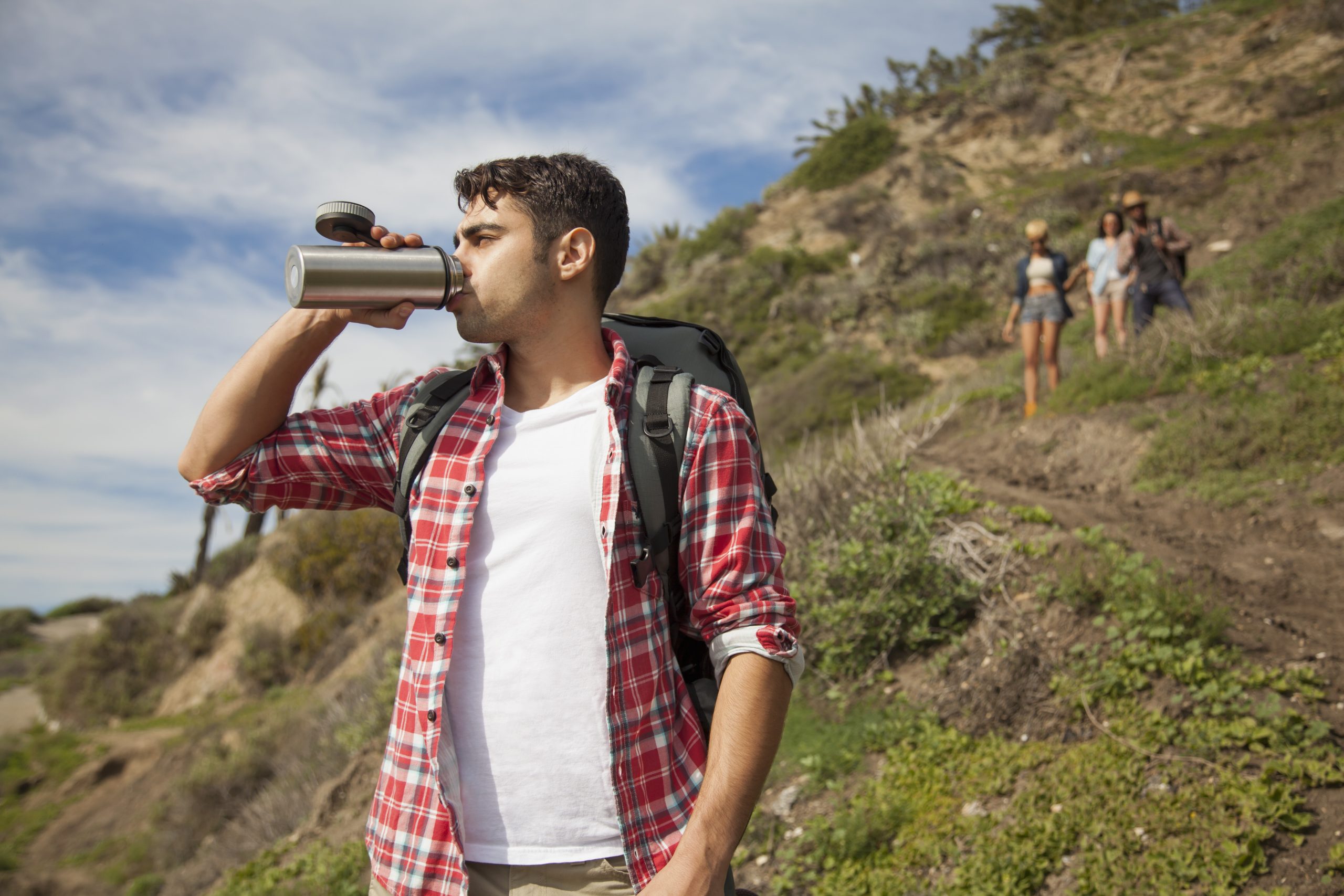 Young man at bottom of hill, drinking from flask, friends trailing behind