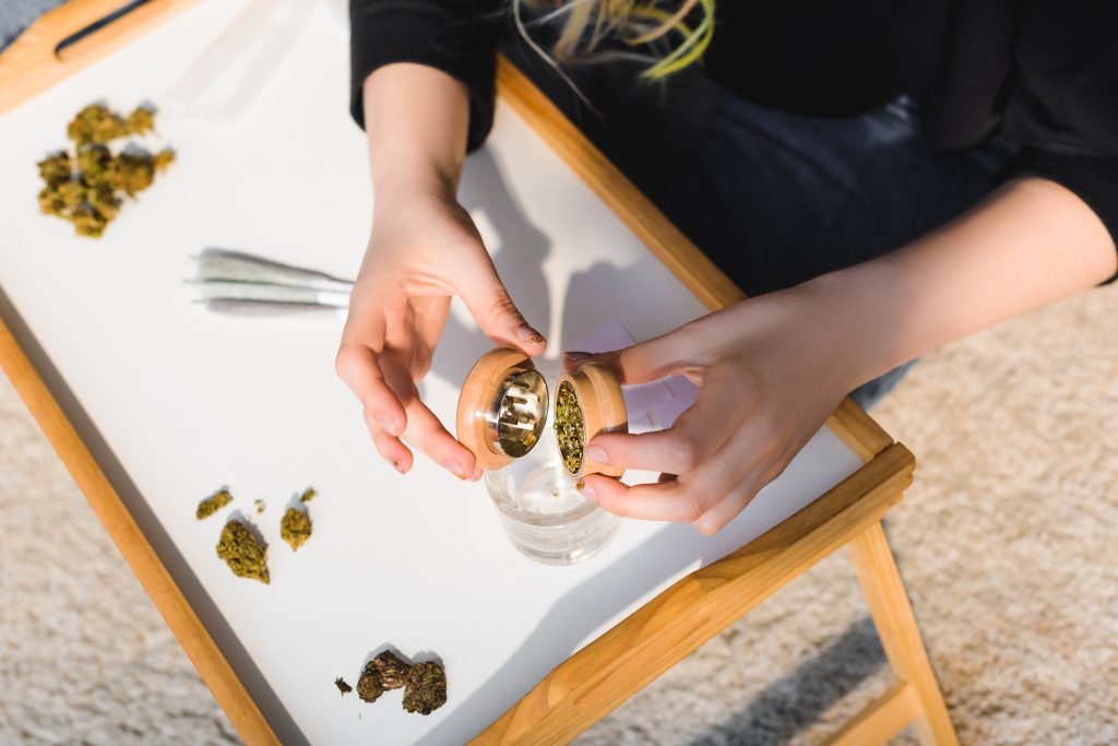 top view of girl putting weed from marijuana grinder in glass on wooden tray